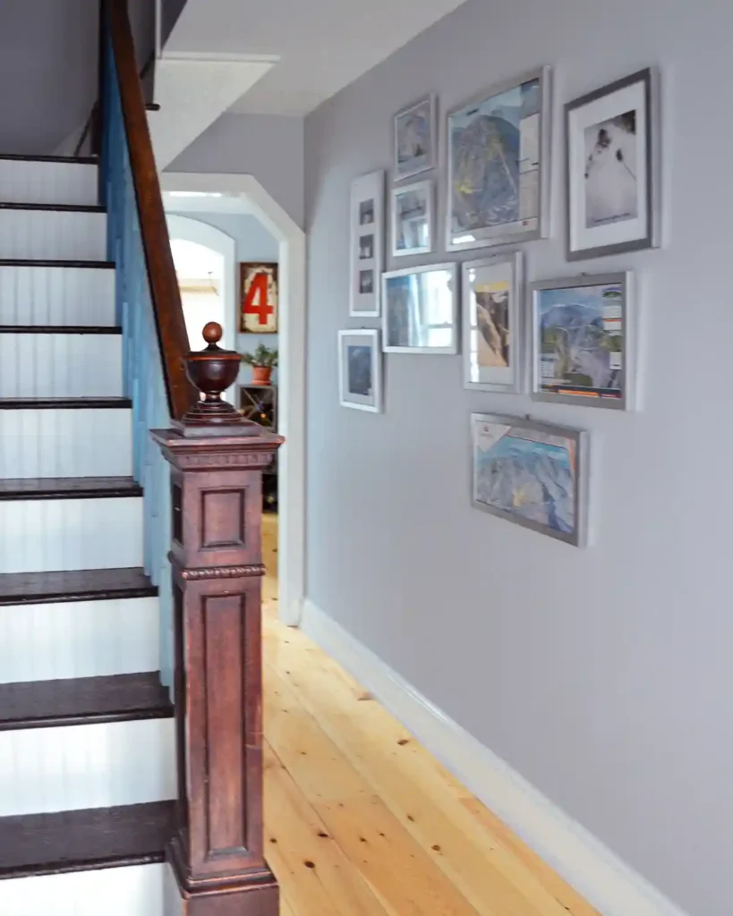 A skiing themed gallery wall hanging in a hallway next to a staircase with silver frames around ski maps, pictures, and other skiing art prints