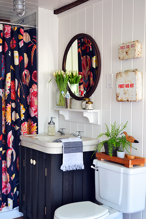 A guest bathroom renovation with a bold floral Anthropologie shower curtain, white planked walls, dark painted vanity, vintage white sink, copper mirror, and vintage first aid kits.