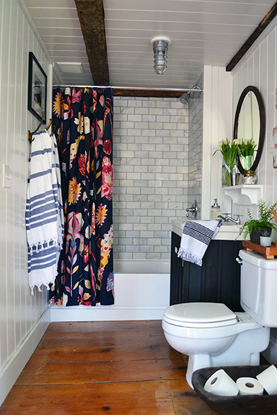 Old farmhouse bathroom renovation with salvaged wide pine floors. Marble subway tile surround is accented with a bright Anthropologie shower curtain. A dark painted vanity base supports a vintage cast iron sink. Turkish bath towels, vintage mirror, buffalo art, and vintage first aid kits accessorize the space.