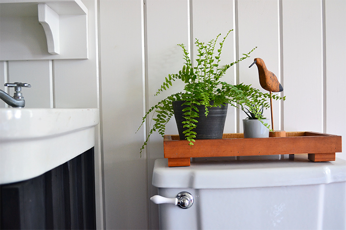 A small wooden tray sits on the back of this farmhouse toilet, holding a fern, small succulent in a blue glass pot, and a wooden sand piper accessory.