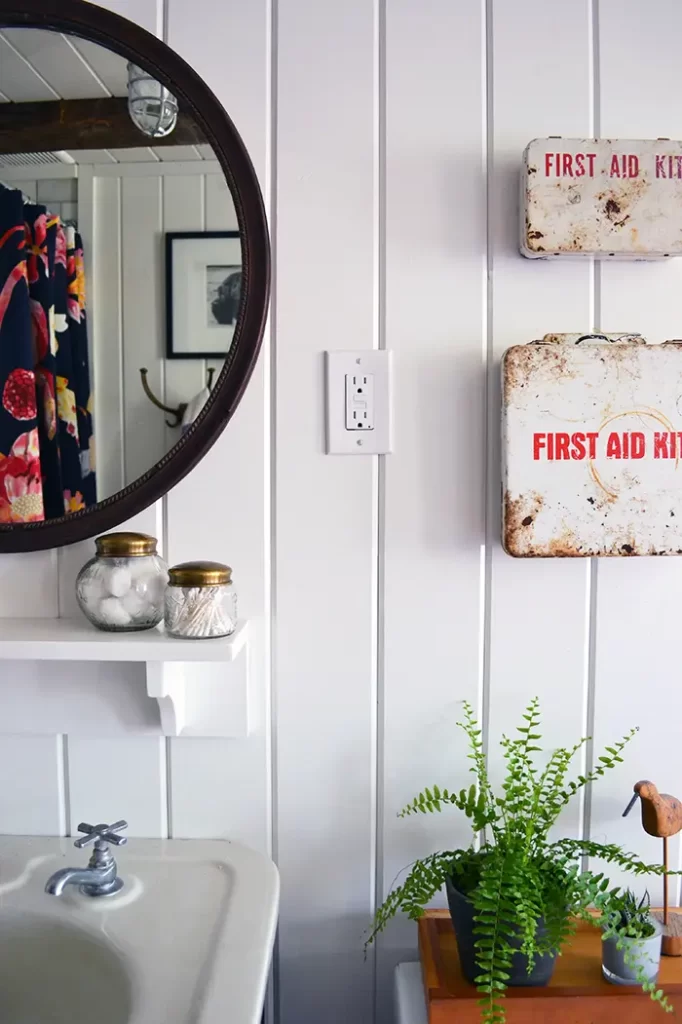 White planked walls in this old farmhouse bathroom renovation with vintage accessories including a flea market mirror, salvage cast iron sink, and old metal first aid kits.