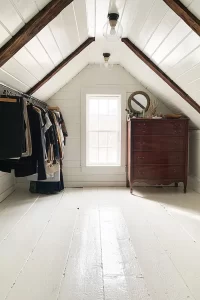 An unfinished attic turned into a finished attic walk-in closet with v-groove planks painted white and hanging rod for clothes under the eaves