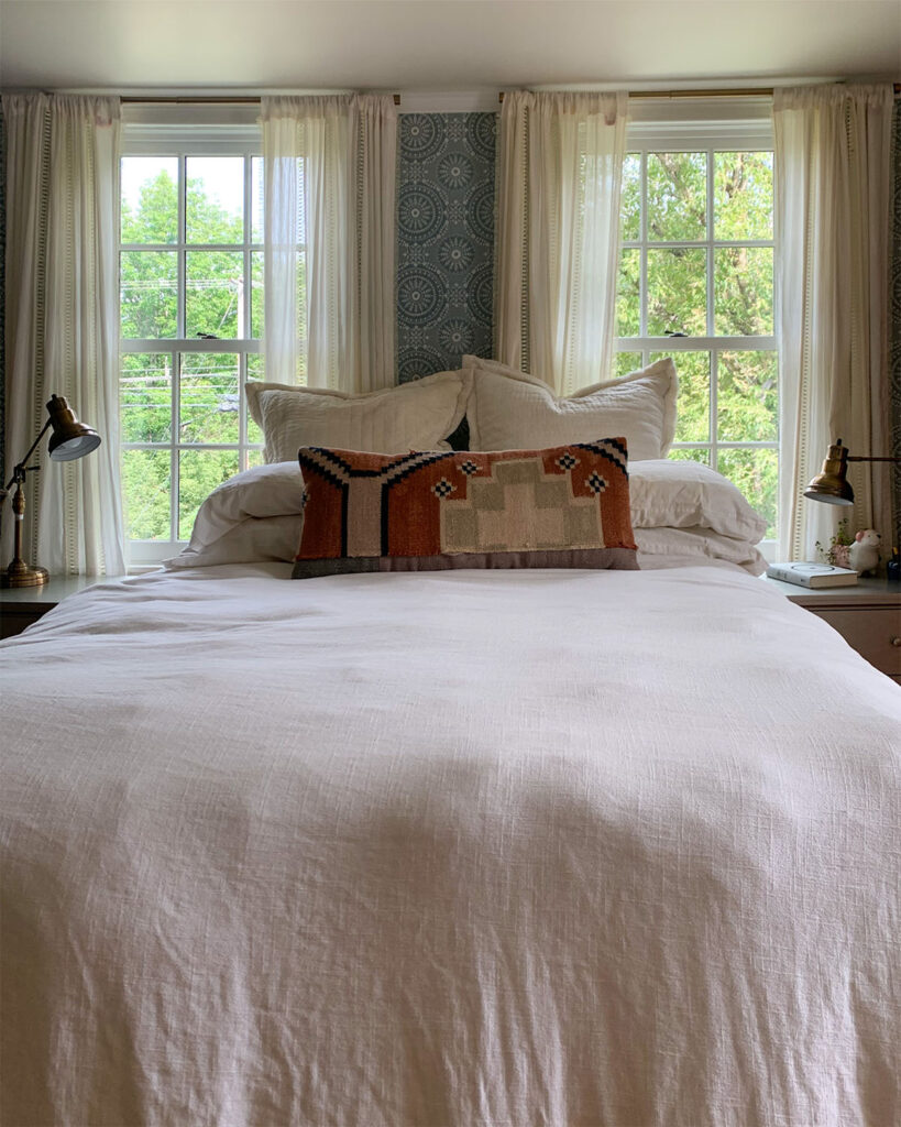 Layers of white bedding in a bedroom in need of a refresh, in front of vintage blue wallpaper