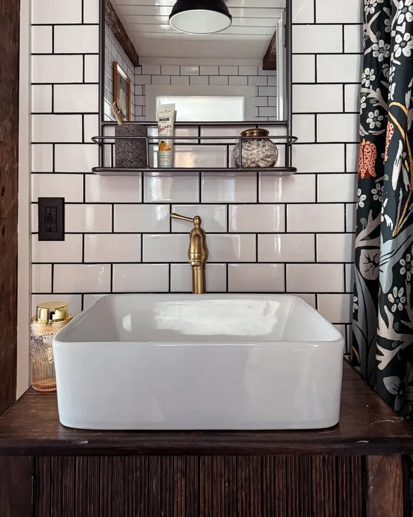 White vessel sink with a gold faucet sitting on top of a dark stained wood vanity against white subway tile with black grout