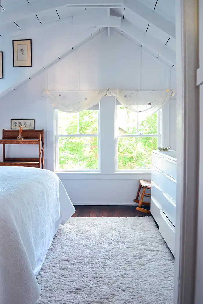 My homebody rich life featured image with a lakehouse bedroom all in white including a lofted ceiling with exposed beams