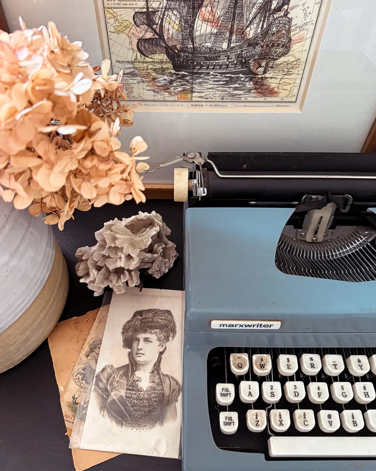 Light blue vintage typewriter on a shelf with dried hydrangeas in a vase, old photos, coral, and ship art