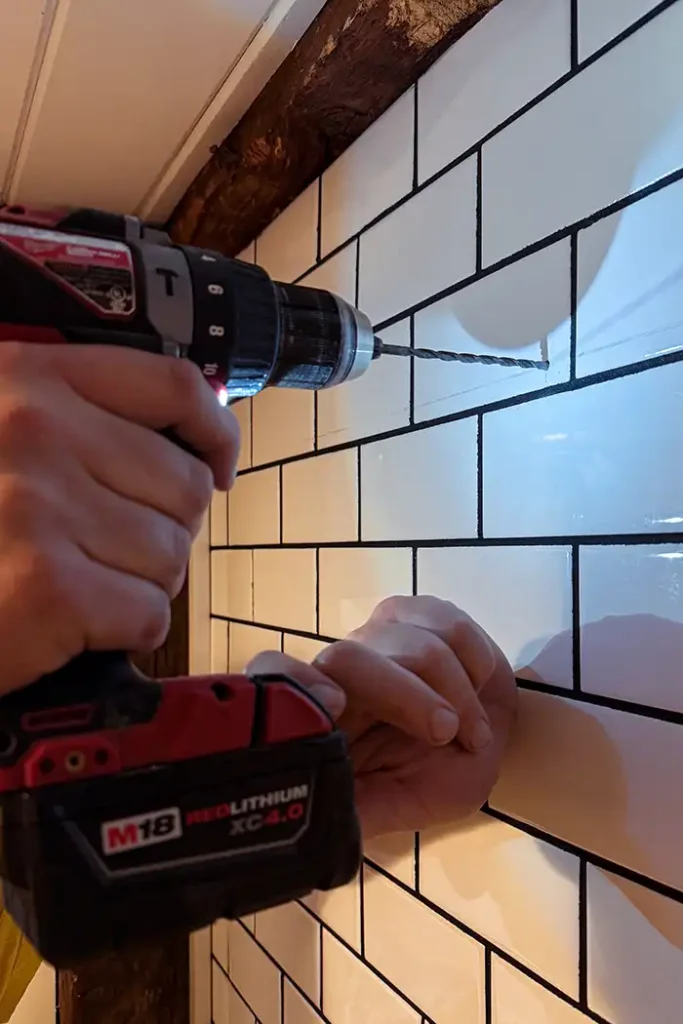 How to drill into bathroom tile featured image a person using a Milwaukee cordless drill with a carbide drill bit starting to drill into white ceramic subway tile installed in a bathroom