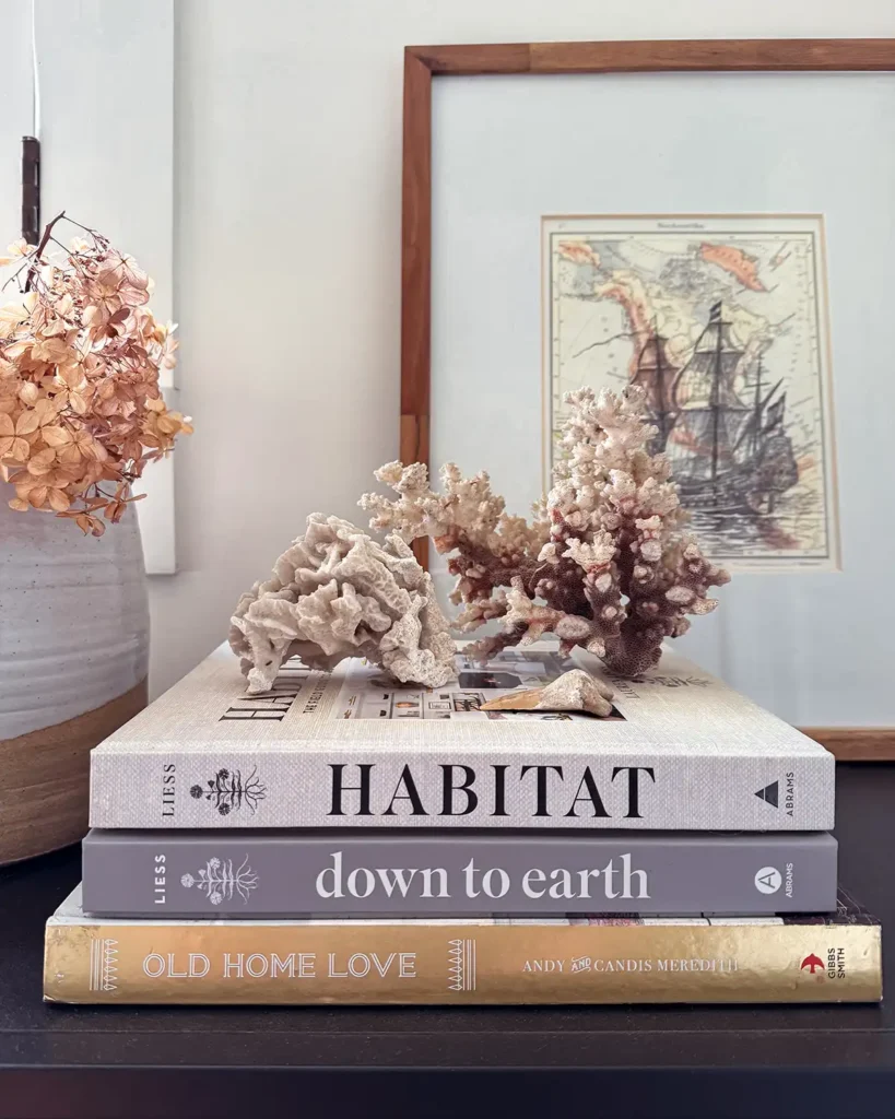 Stack of my my favorite design books including Habitat by Lauren Liess, Down to Earth by Lauren Liess, and Old Home Love by Candis and Andy Meredith. Styled on a shelf with coral, ship art, and a vase of dried flowers.