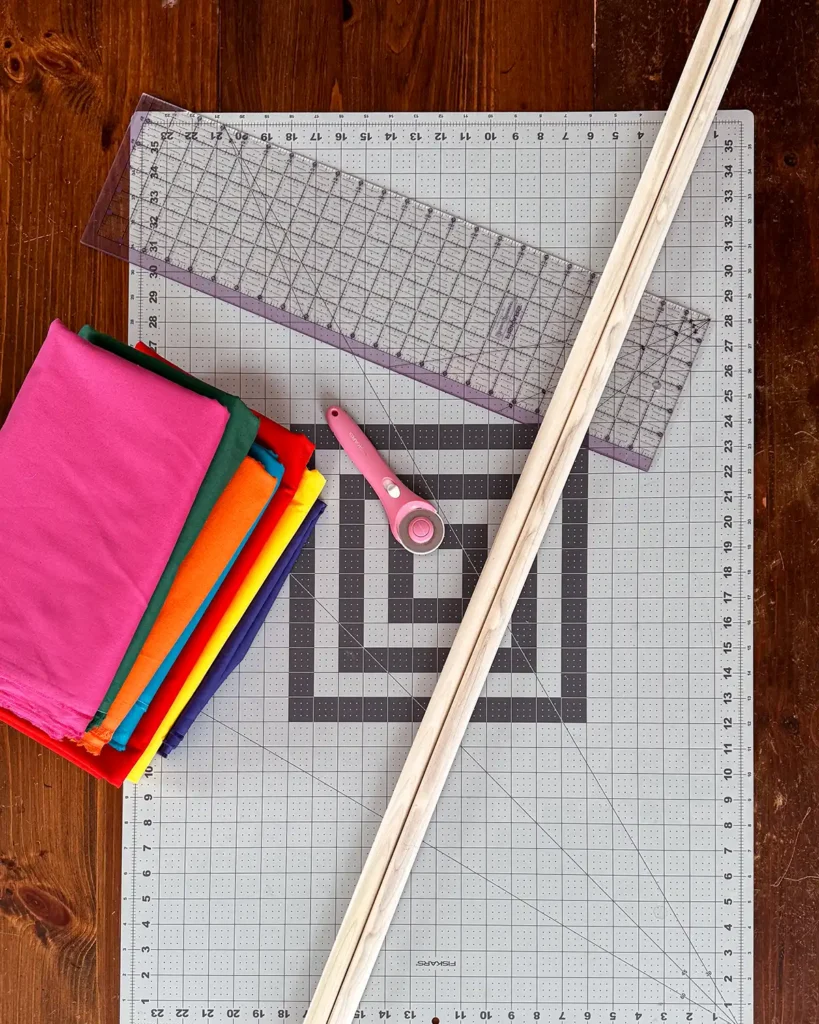 Birthday backdrop making supplies including a stack of colorful fabrics, fabric cutting mat, sewing ruler, rotary cutter, and rotary cutter