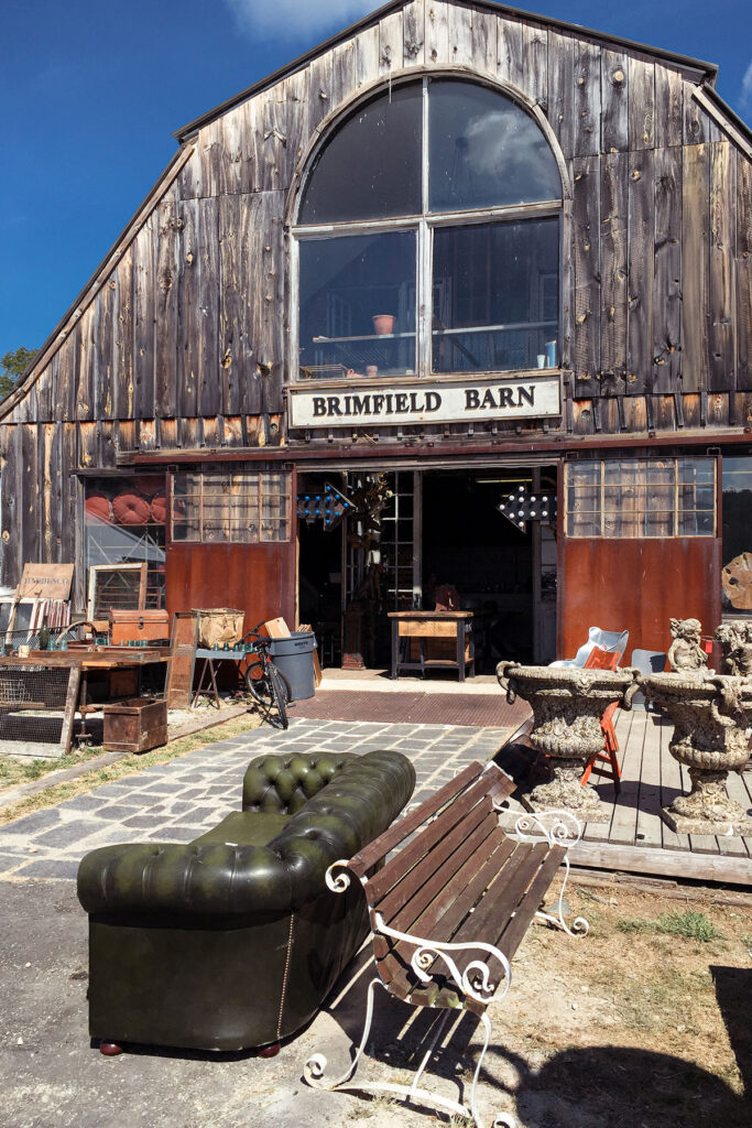 Brimfield Flea Market guide featured image with a photo of the Brimfield Barn and many antique and vintage items in front of the barn