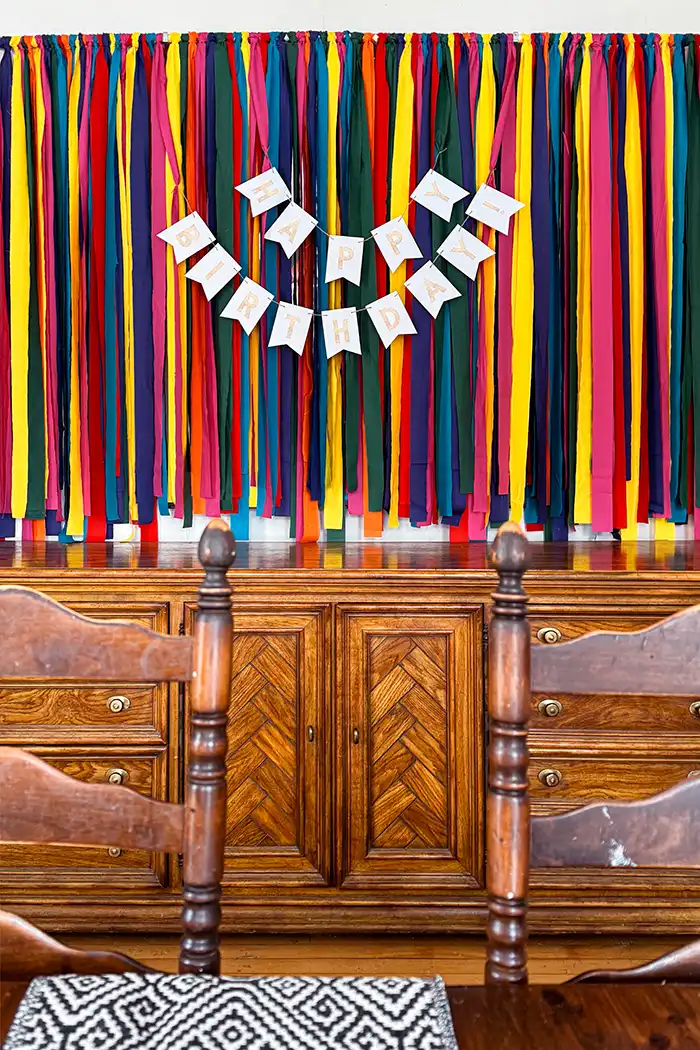 DIY birthday backdrop featured image with a backdrop made with colorful fabric strips tied to a dowel and hung on the wall, hanging above a console table in a dining room