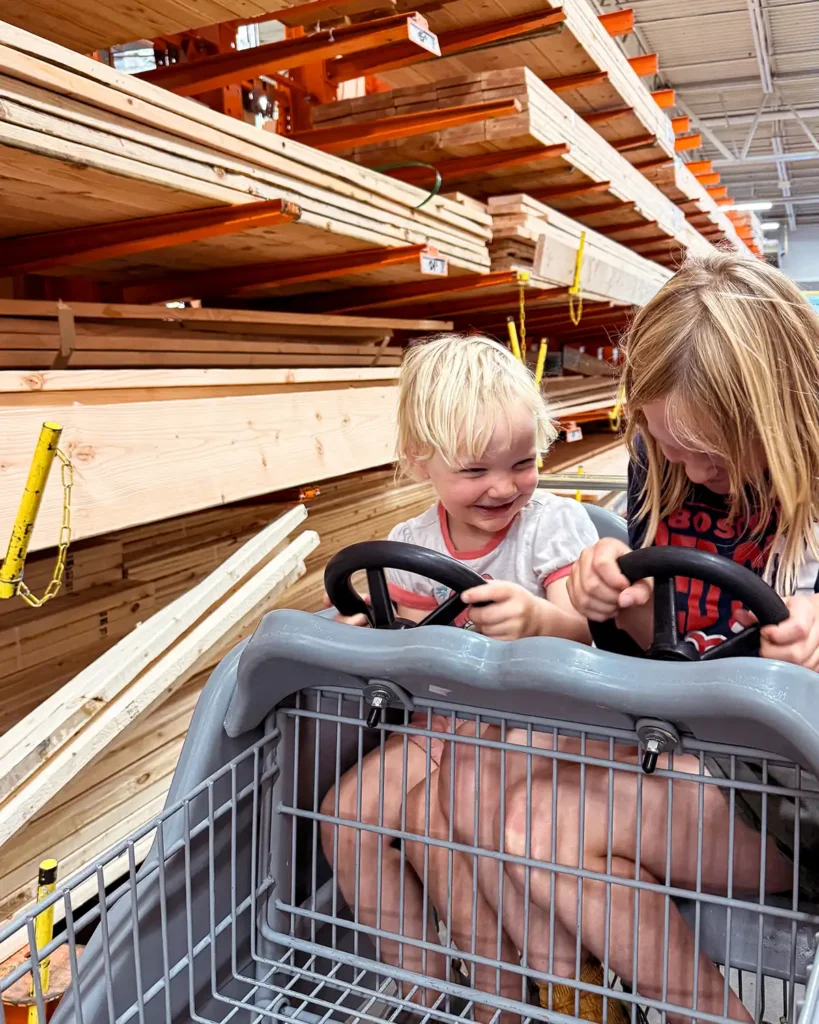 Two kids driving a Home Depot race cart through the lumber aisle