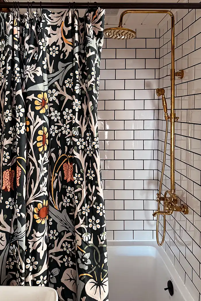 Primary bathroom sources featured image with a dark floral shower curtain hanging in front of a white subway tile shower with black grout and a gold shower system