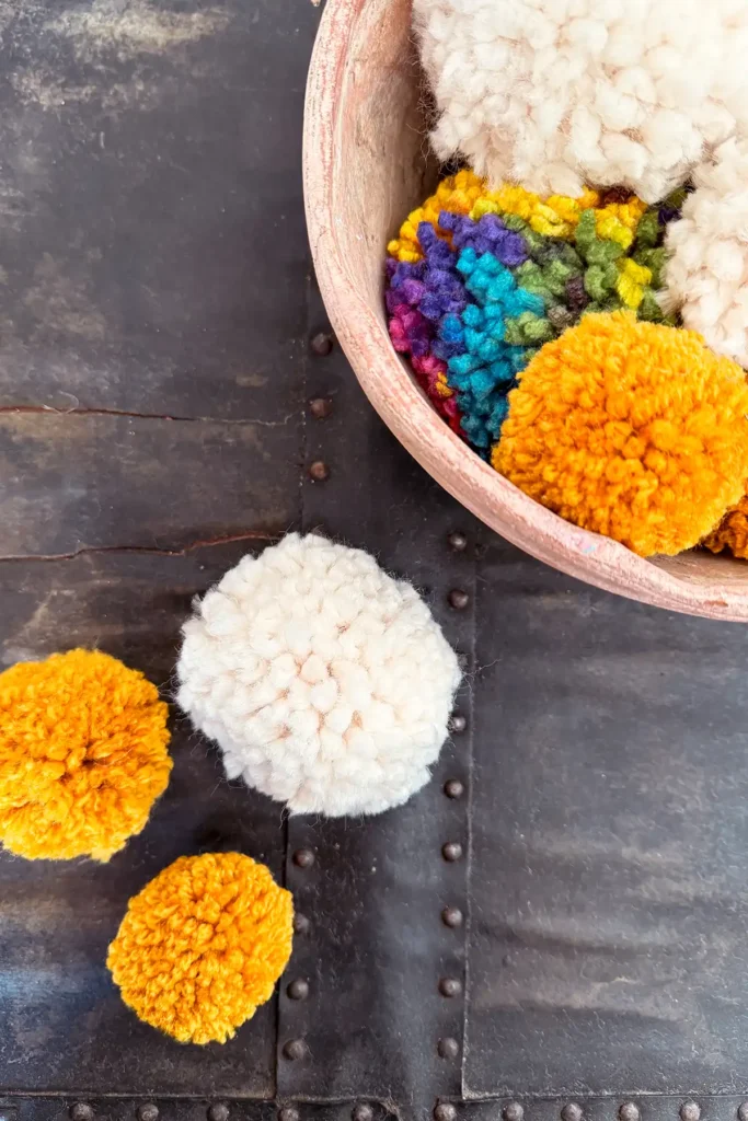 How to make a pom pom using a pom pom maker featured image with a pile of poms poms in white, mustard yellow and variegated sitting in a clay bowl and on a vintage black surface