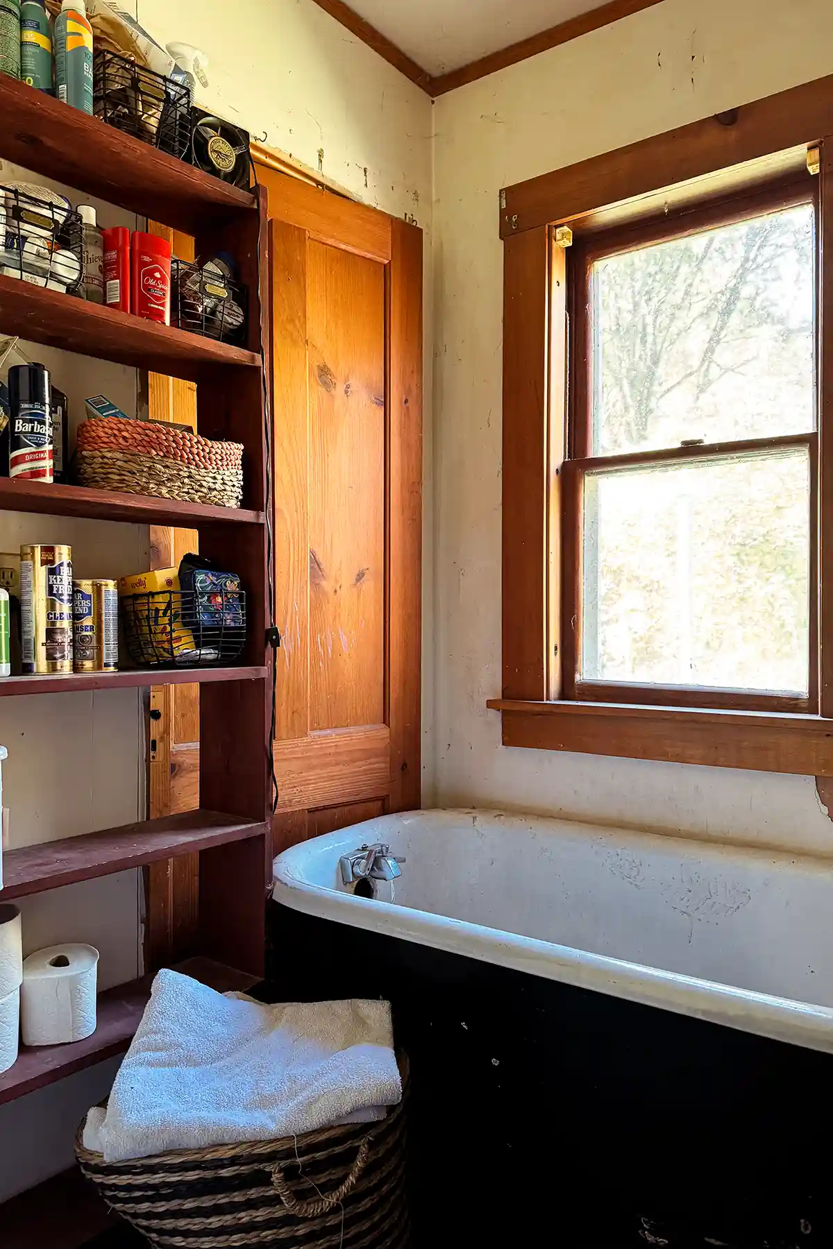 A small hallway at the end of bathroom closed up and ready to become a small laundry room; holds a black vintage clawfoot tub and small bookcase filled with toiletries, medicines, cleaning supplies, extra toilet paper and more