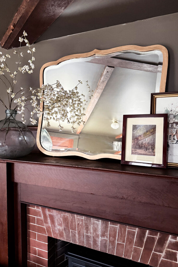 A DIY mirror frame makeover with gold Rub N Buff, mirror hanging above a fireplace with a vase of flowers and a pair of framed art pieces layered around it on the mantel