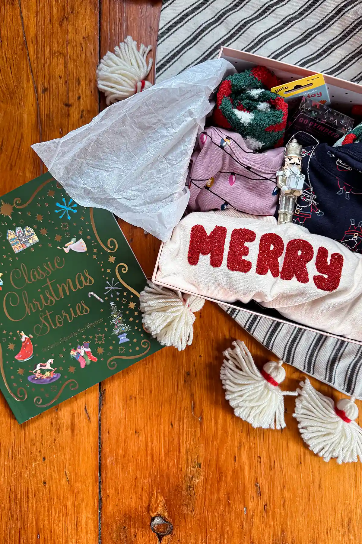 December 1st box with a Classic Christmas Stories box, Merry sweatshirt, pagamas, card game, Yoto card, and fuzzy holiday socks in a pink gift box on a tree skirt