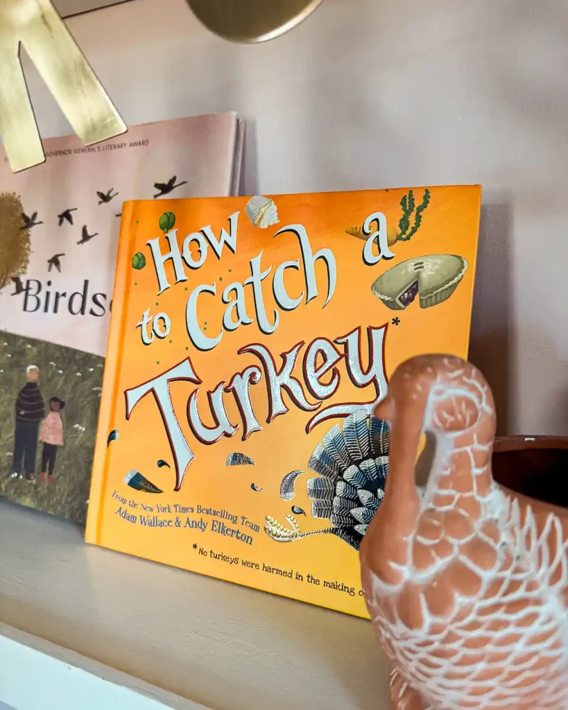 How To Catch A Turkey, a silly kids picture book to read together at Thanksgiving, sitting on a children's Thanksgiving themed bookshelf with a ceramic turkey