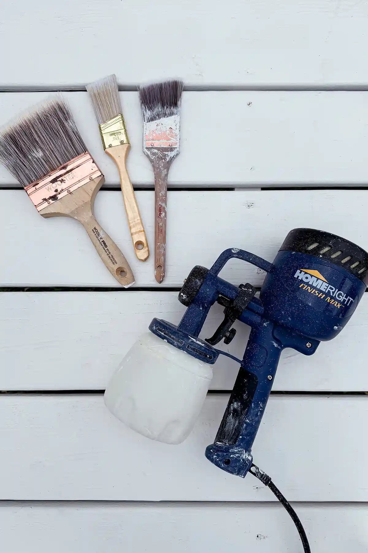 A trio of paintbrushes in a variety of sizes next to a HomeRight Finish Max paint sprayer sitting on white painted playground decking