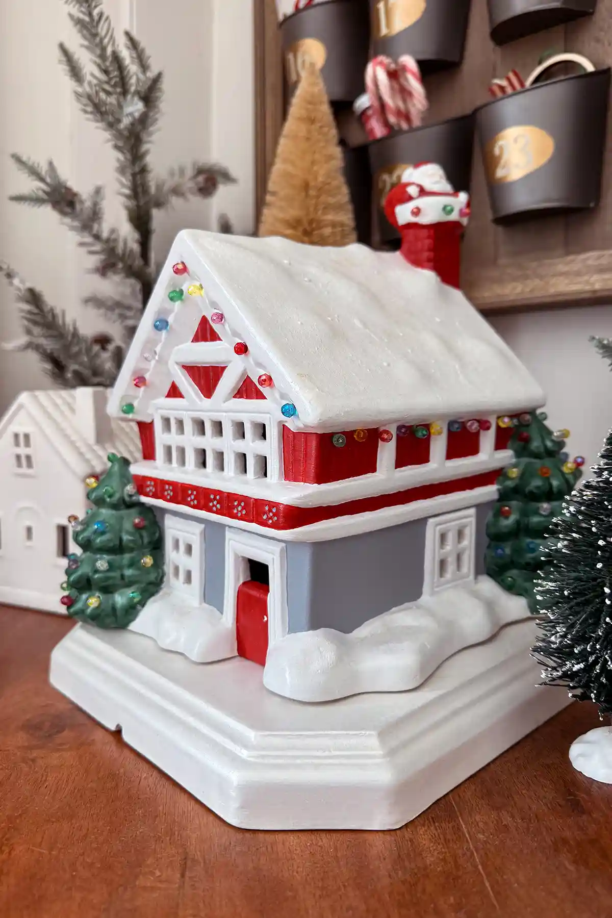 A ceramic Christmas house that's hand-painted in red, green, white, and gray with Santa coming down the chimney; house is sitting on a board surrounded by bottle brush trees and white ceramic houses from Target