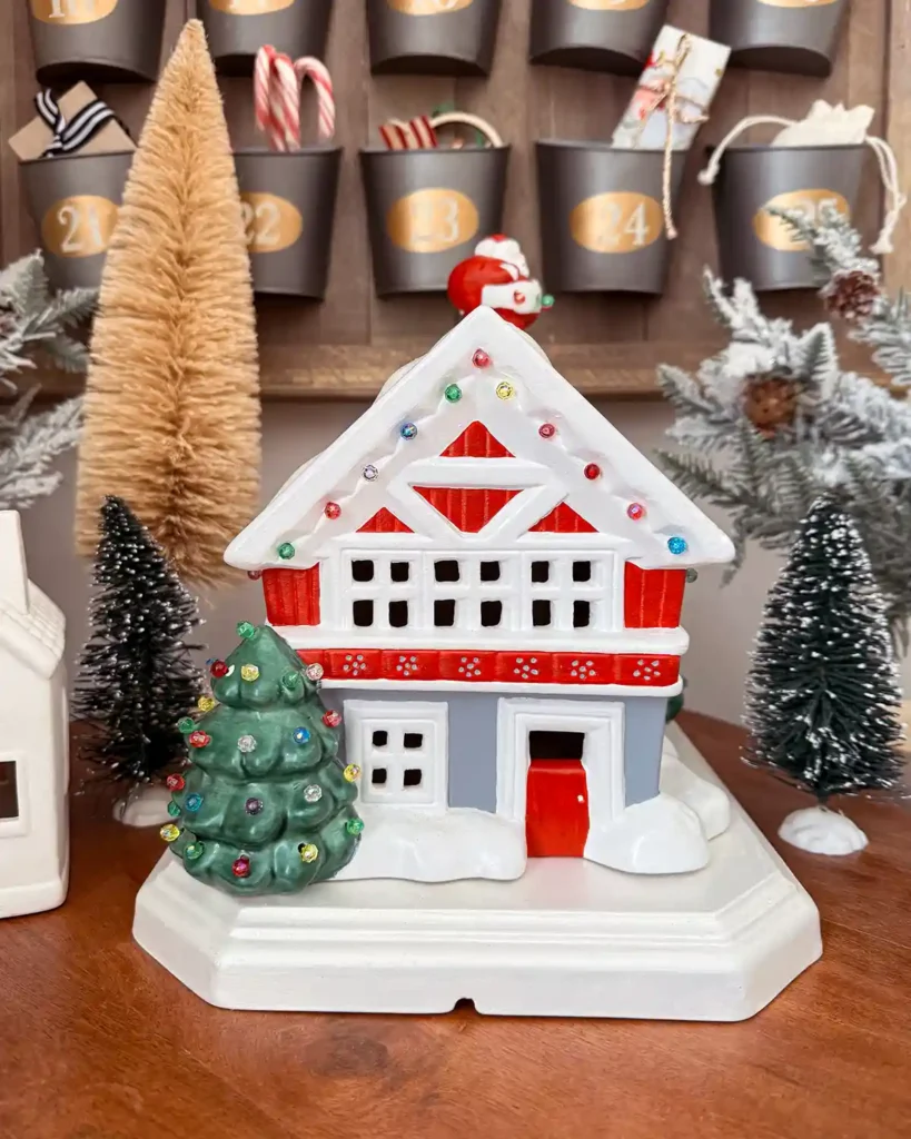 A vintage ceramic Christmas house with a tree out front with lights on it and Santa coming down the chimney, surrounded by bottle brush trees with an Advent calendar in the background
