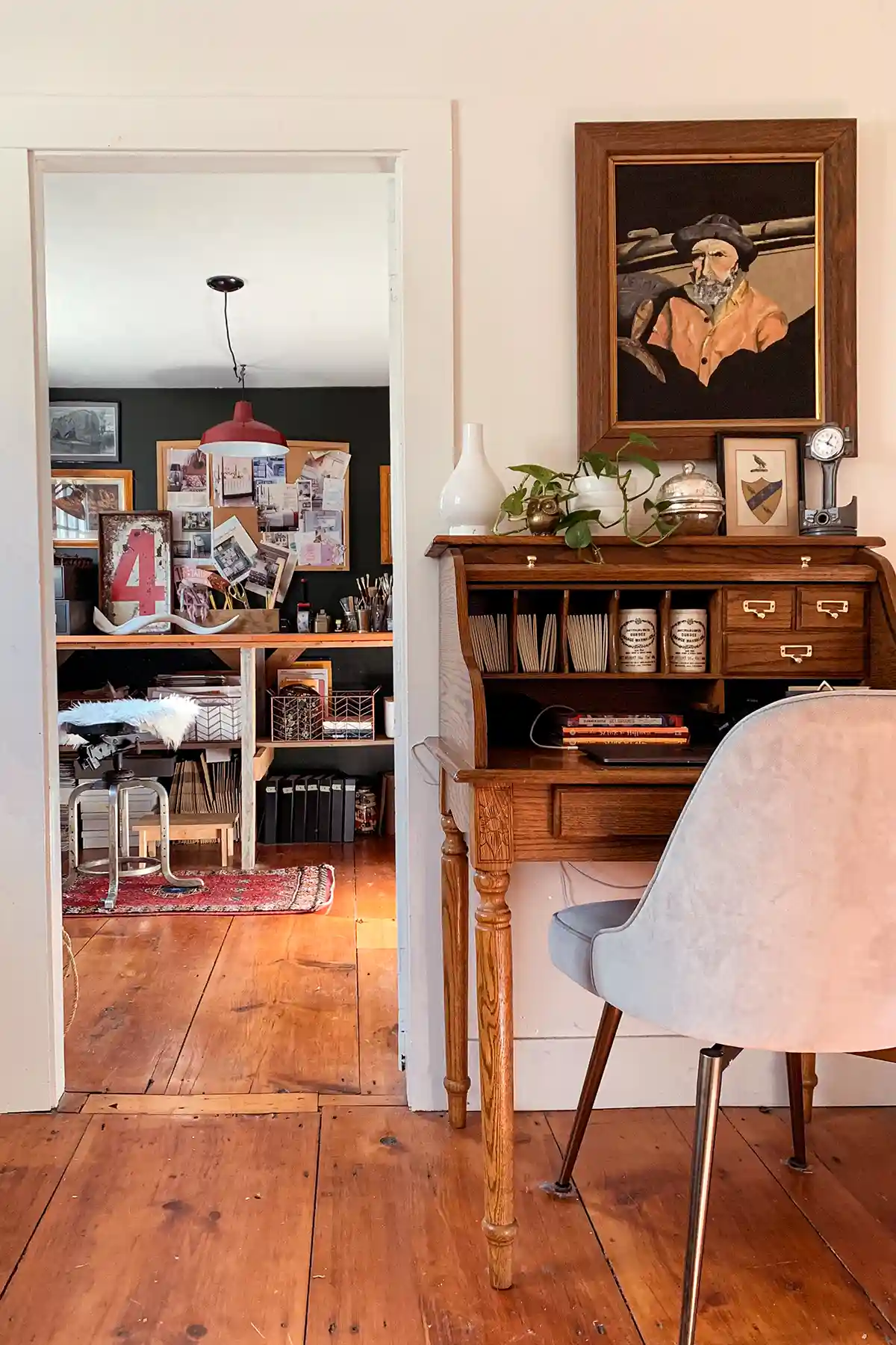 Craft room with a large wall to wall standing desk and dark painted walls peeking through the doorway with a small, wooden rolltop desk next to the door