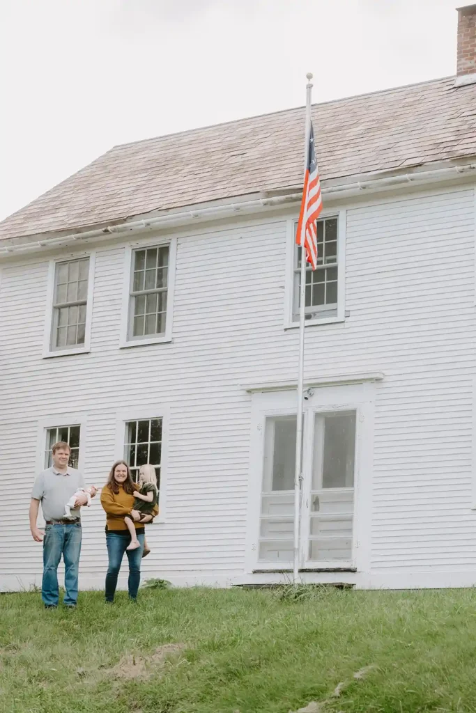 A vintage 1700s white colonial house with a slate roof, flag pole in front, and a family of four in front of it