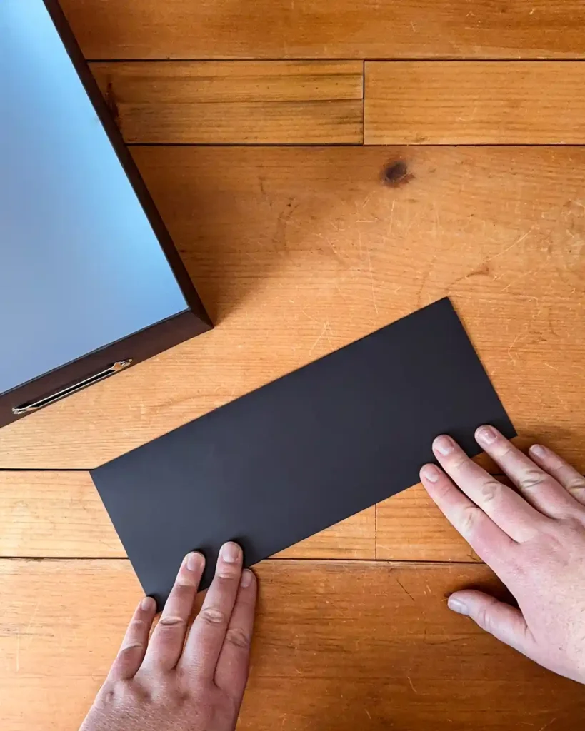 Folding a piece of black card stock in half the long way on a wooden desk surface; an open box sits in the background