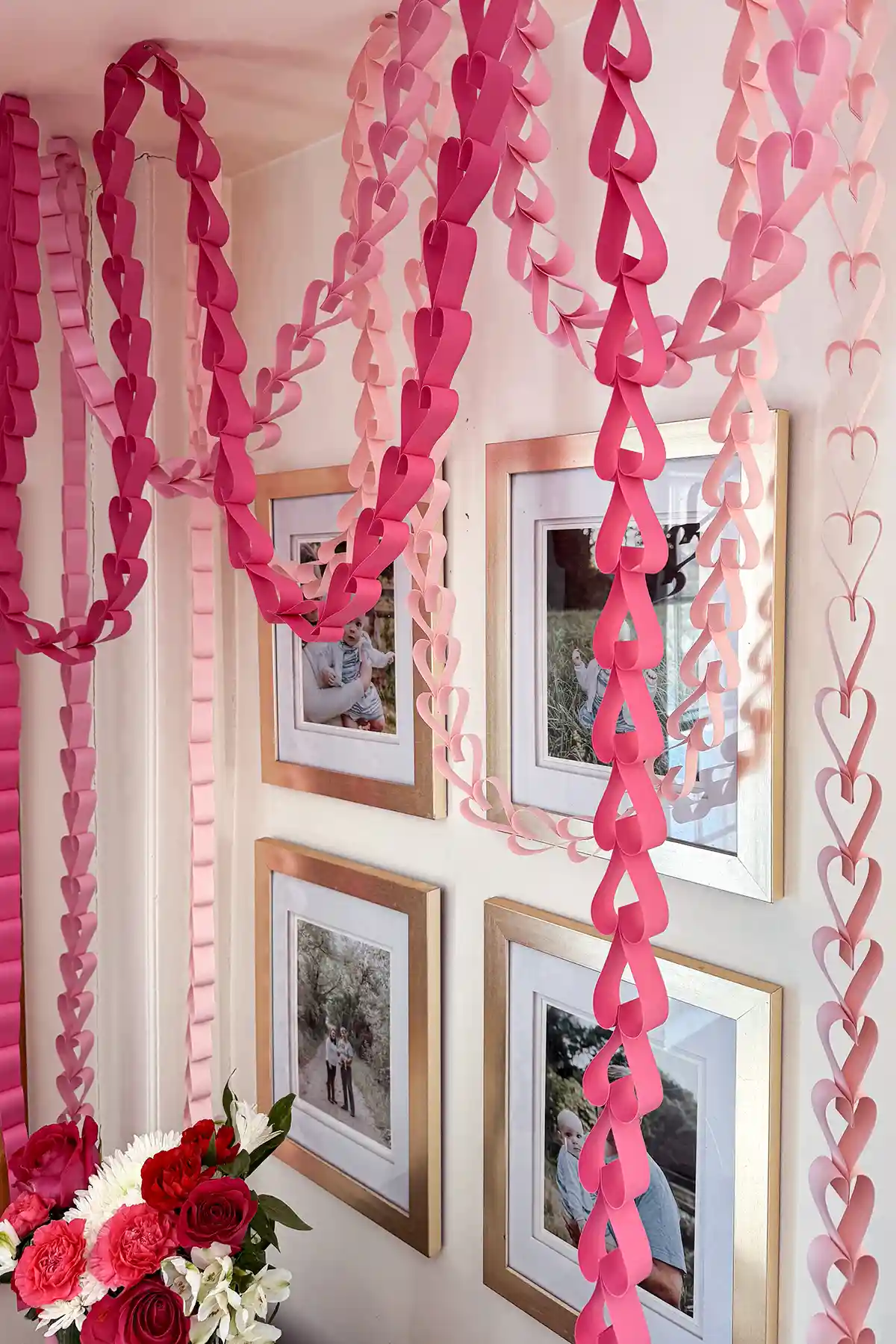 Tutorial for how to make paper heart chains like this trio of three chains in various shades of pink hanging from the ceiling in front of a family photo gallery wall