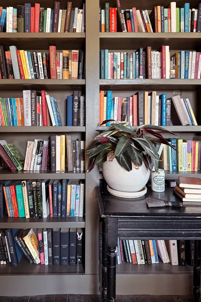 Dark green bookshelves after decluttering books still filled with fiction books; small black table in front of the shelves with a plant and small stack of books on it