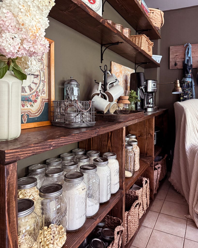 New kitchen pantry shelves including a long, low bookcase filled with dry goods and open upper shelves with lesser used kitchen items like cake plates and a scale