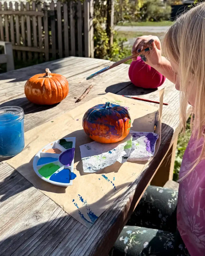 A child sitting at a table painting a small pumpkin with blue paint