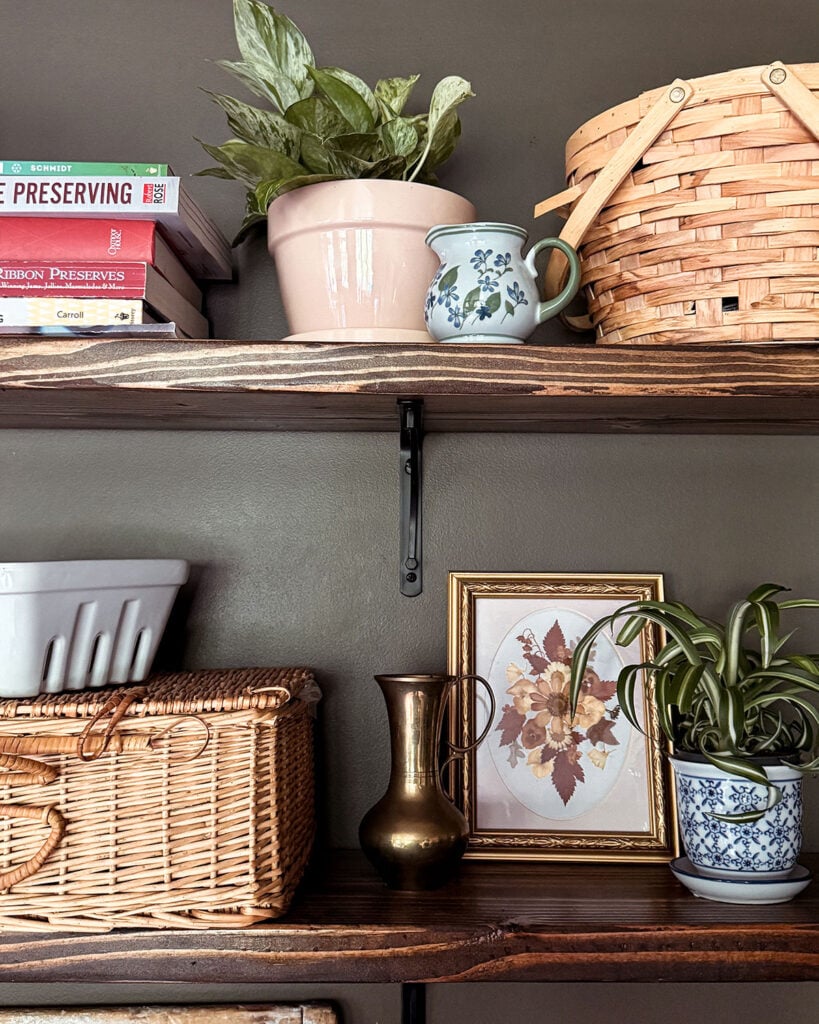 Simple open shelves in a kitchen made from dark stained wood, black brackets, with cookbooks, plants, baskets, and more sitting on the shelves