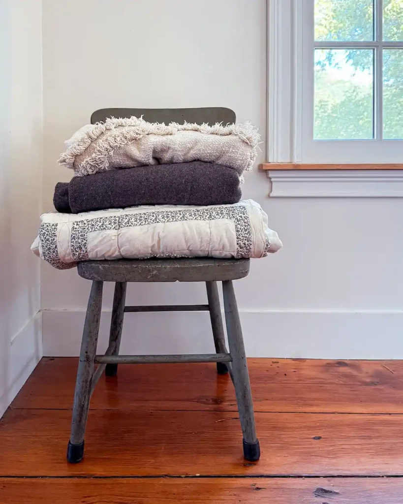 A stack of cozy blankets sitting on top of a chippy moss green chair in a hallway in front of a window