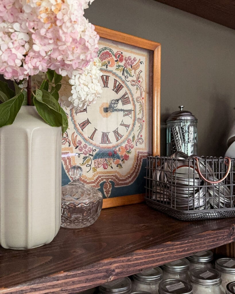 Vintage decor on kitchen pantry shelves including a vase of hydrangeas, cross stitch clock, crystal candy jar, and more