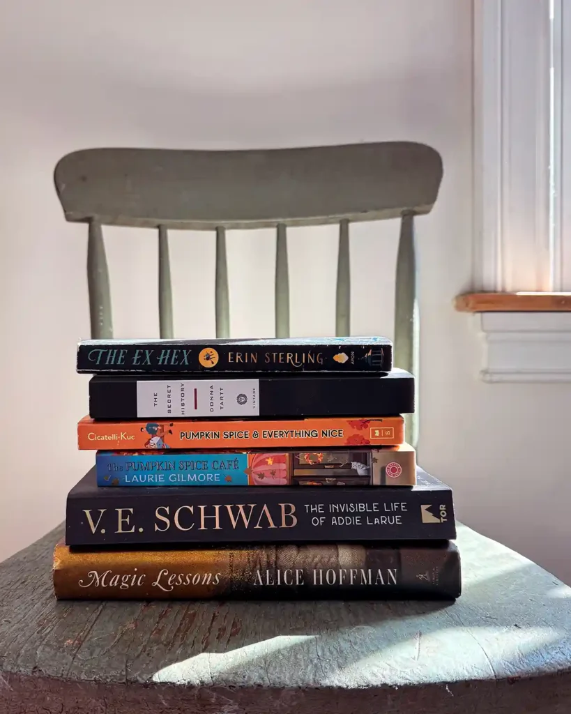 A stack of fiction books for fall sitting on top of a chippy, sage green chair, including titles: The Ex Hex by Erin Sterling, The Secret History by Donna Tart, Pumpkin Spice & Everything Nice by Cicatelli-Kuc, The Pumpkin Spice Cafe by Laurie Gilmore, The Invisible Life of Addie LaRue by V.E. Schwab, and Magic Lessons by Alice Hoffman