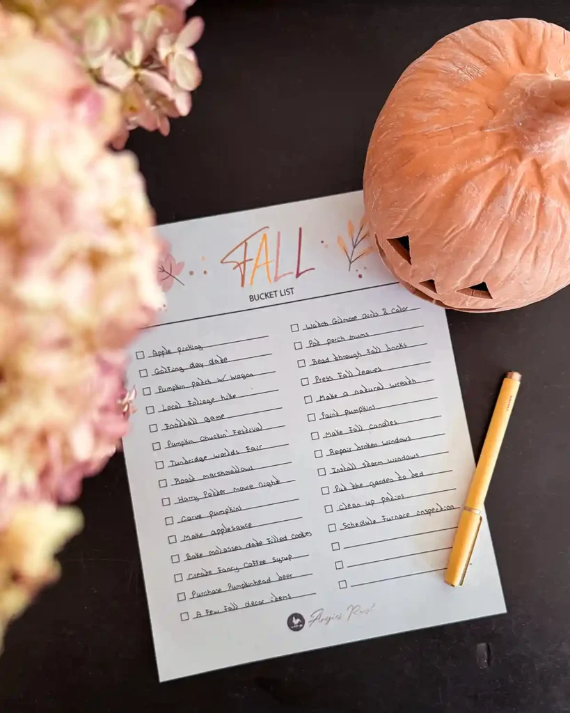 A printable fall bucket list sitting on a table with a yellow pen filled out for a family; ceramic jack-o-lantern and dried hydrangeas sitting next to it