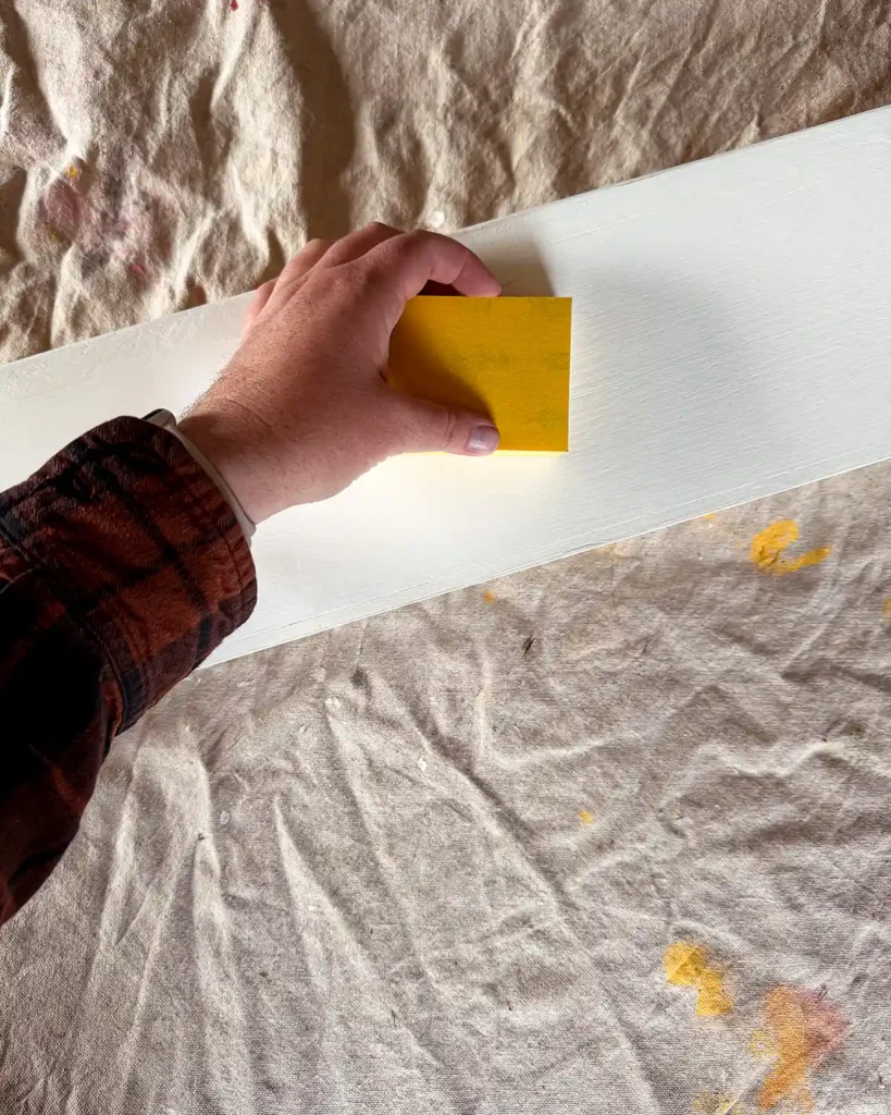 Using a fine sandpaper block to sand between coats of paint on a glossy white shelf