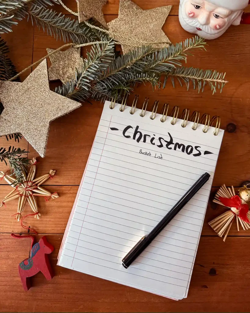 A notepad with "Christmas bucket list" written on it accompanied by a pen, some fresh balsam fir greens, Christmas ornaments, glittery stars, and a Santa mug