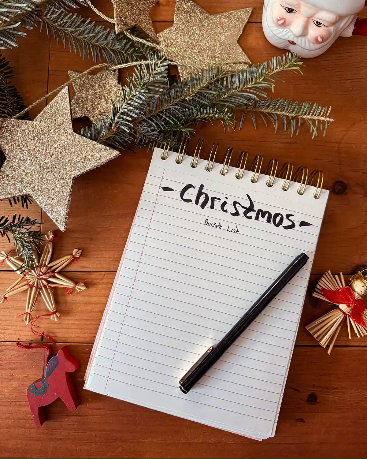 A notepad with "Christmas bucket list" written on it accompanied by a pen, some fresh balsam fir greens, Christmas ornaments, glittery stars, and a Santa mug