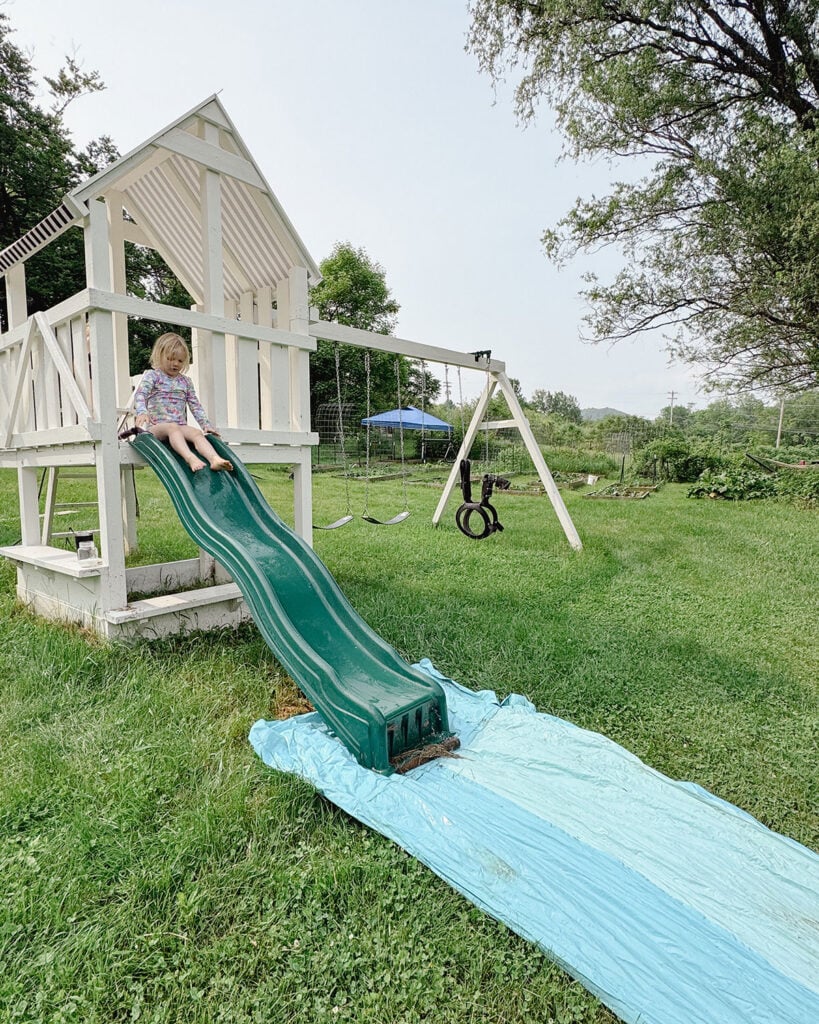 A white painted kids playground with a child going down the slide and onto a slip and slide with a large, kitchen garden behind it