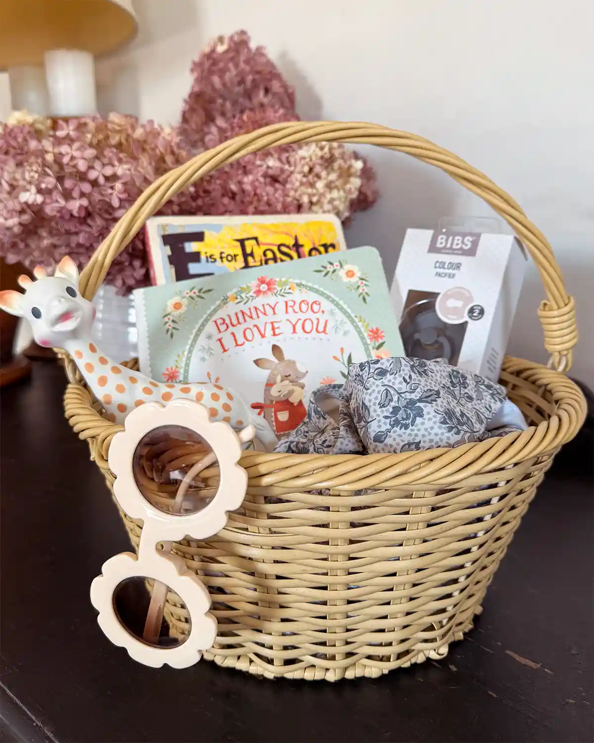 A simple wicker Easter basket for a baby filled with a Sophie the Giraffe teether, E is for Easter board book, Bunny Roo I Love You board book, box of pacifiers, light blue bathing suit, and a pair of flower shaped sunglasses