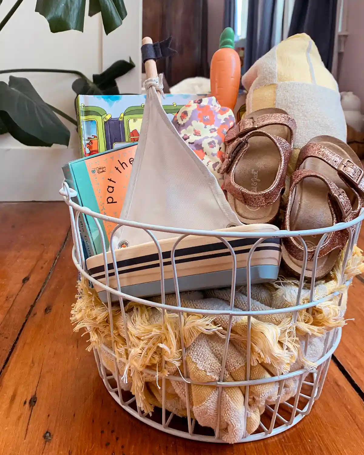 A toddler's Easter basket with a beach towel, bathing suit, bubbles, search and find book, Pat the Bunny book, glittery gold sandals, and a wooden bath toy boat