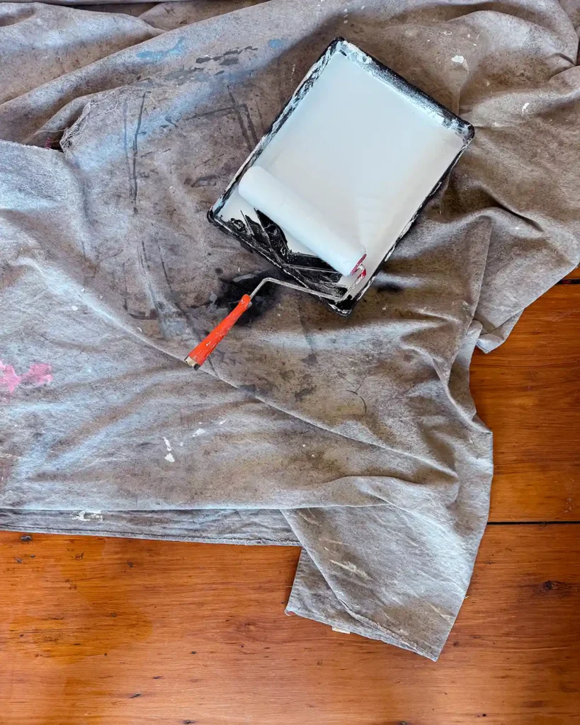 A roller sitting in a paint tray that's full of white primer. The tray is sitting on top of a paint-stained drop cloth on the floor