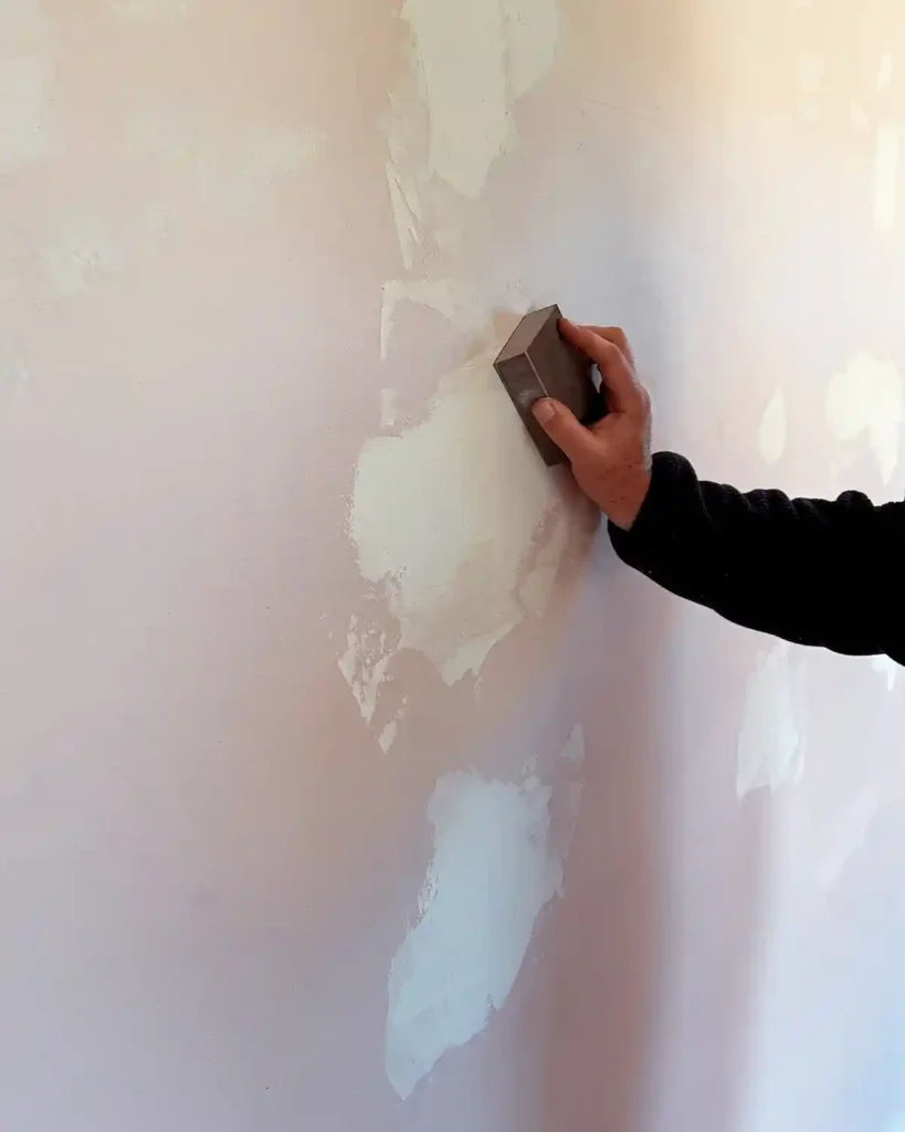 Sanding dried joint compound covering repaired drywall holes using a fine-grit sanding block