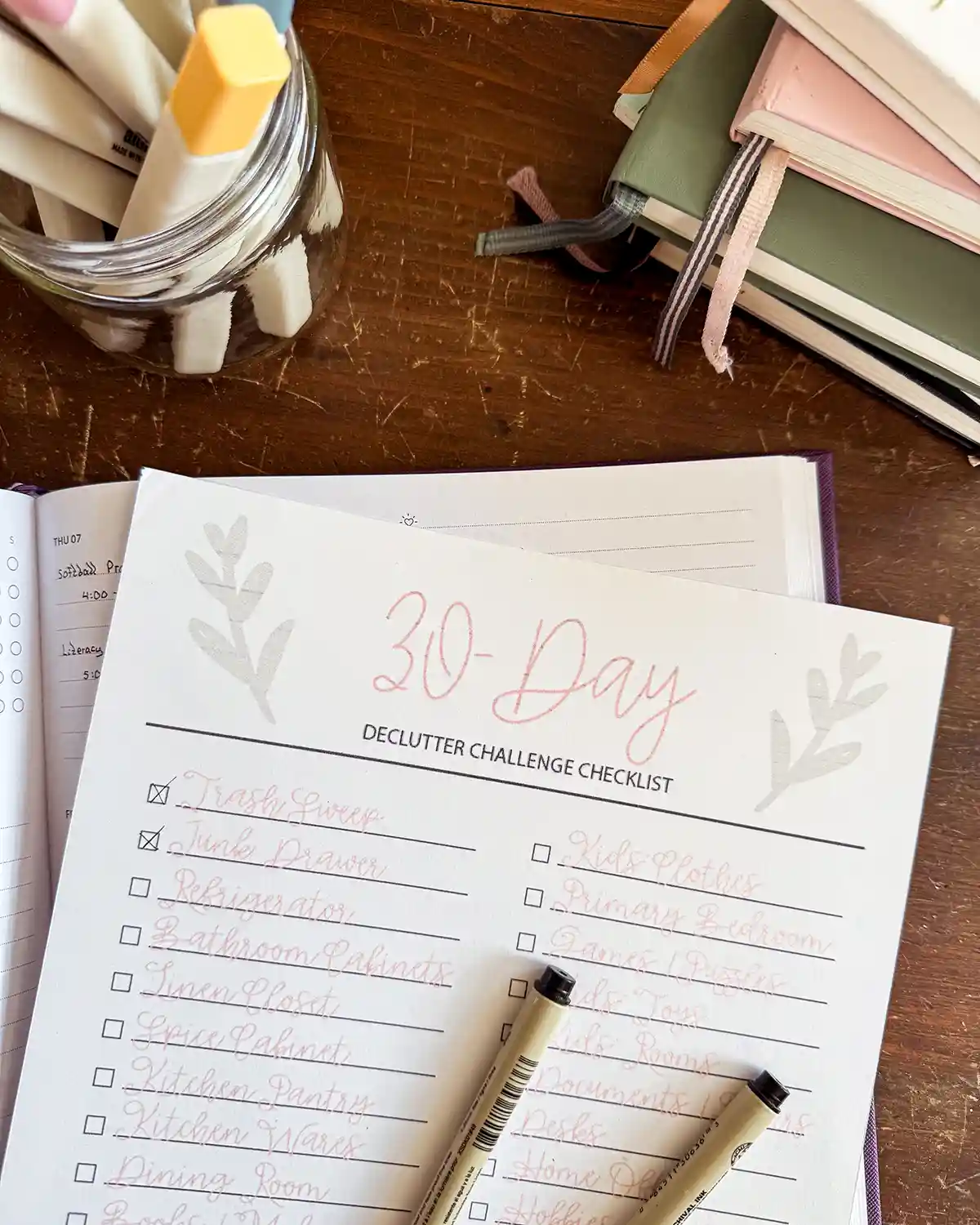 A 30-day declutter challenge printed PDF checklist sitting on top of a planner with pens, highlighters and notebooks sitting nearby