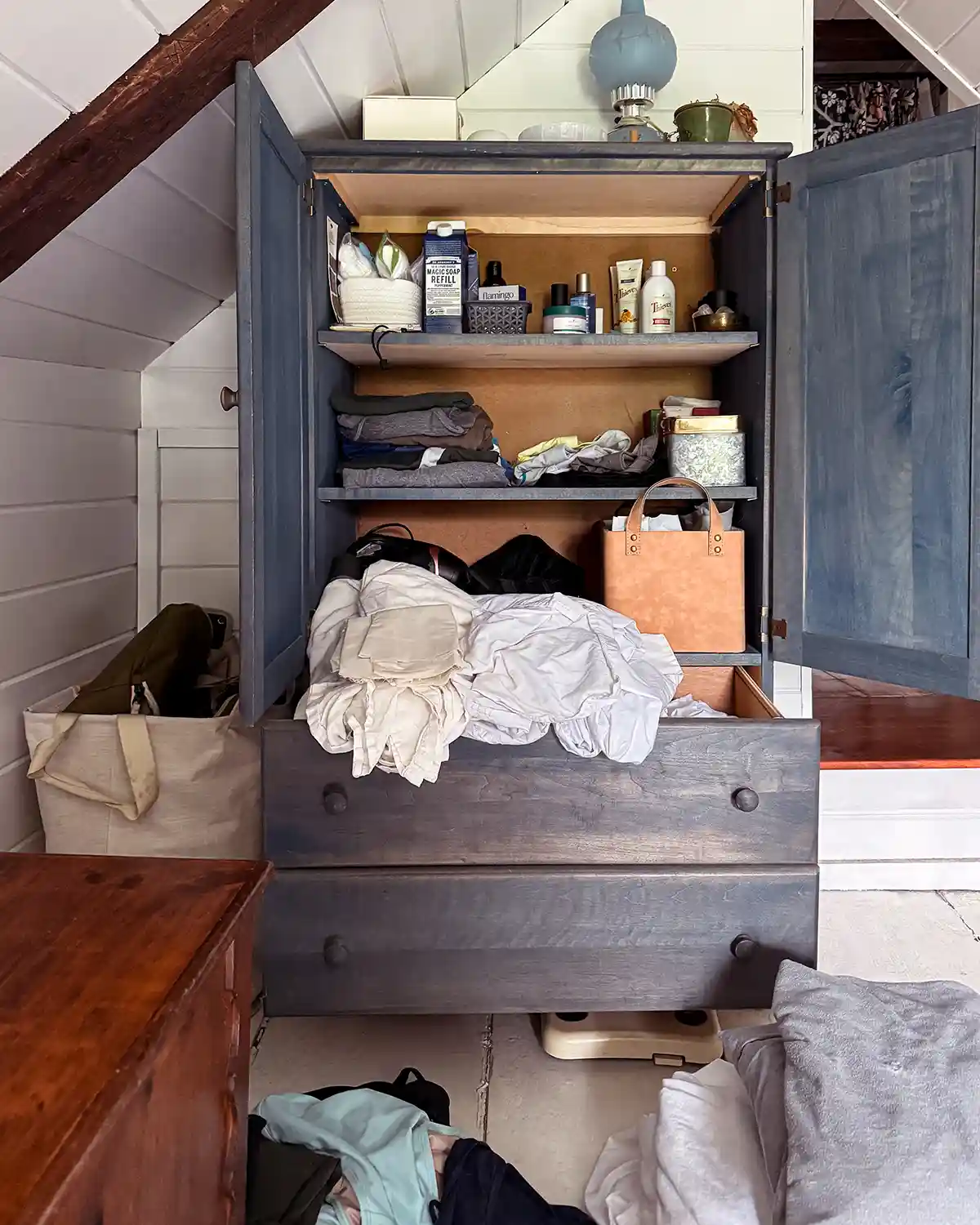 A blue, super cluttered cabinet used for bathroom and linen storage, with piles of sheets spilling out of the drawers and the cabinet doors open revealing all kinds of bathroom related items stored within it