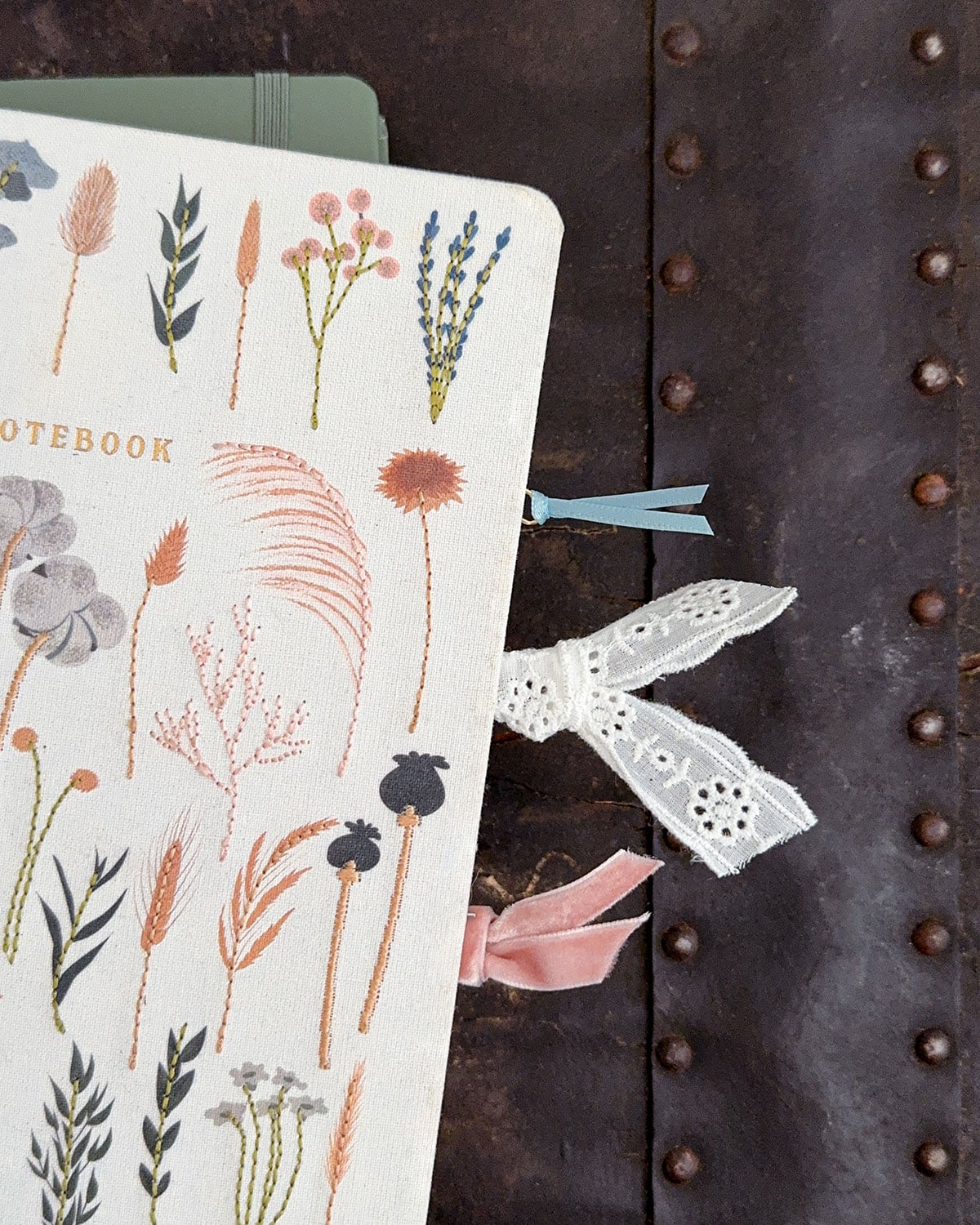 Three DIY ribbon and paperclip bookmarks clipped to a notebook with the ribbon part sticking out; one blue ribbon, one white eyelet, and one pink velvet