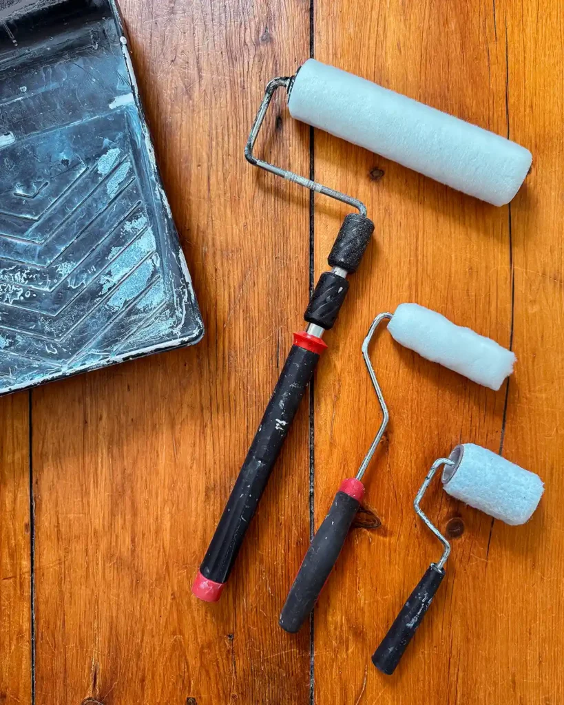Different types and sizes of paint rollers sitting on a floor beside a paint tray