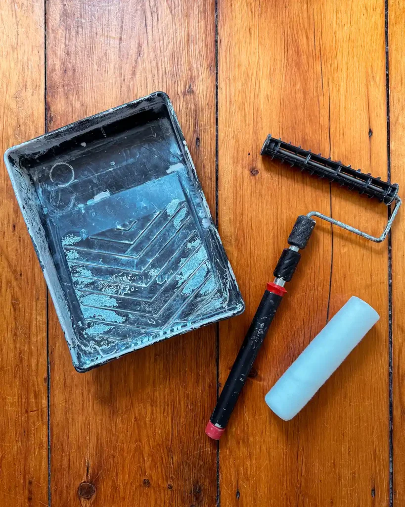 An extendable paint roller sitting on a wood floor beside a paint tray and synthetic roller cover
